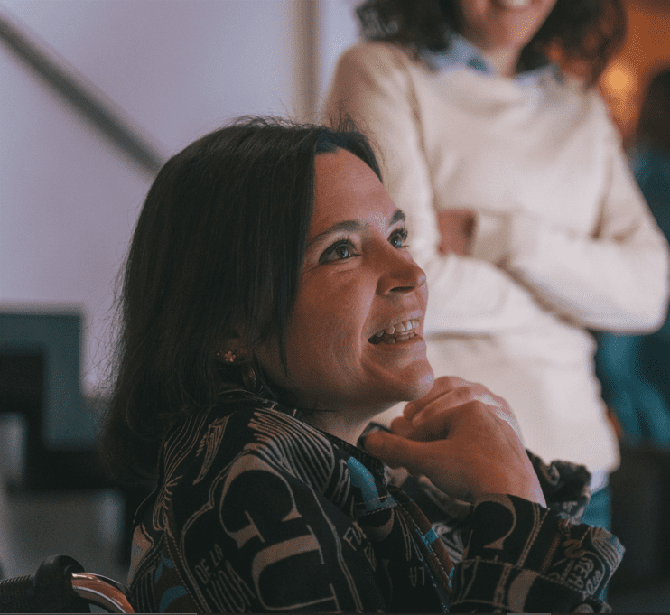 Grupo de cinco personas sentadas en un sofá en un ambiente profesional. Una mujer sonríe mientras sostiene una taza pequeña, mientras otra revisa su teléfono. Al fondo, dos personas conversan, destacando la vista de la ciudad a través de la ventana. En la mesa hay una cafetera y varias tazas, lo que sugiere un momento de trabajo o reunión informal.
